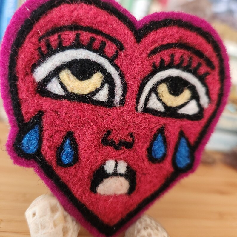 May include: A heart-shaped felt patch in vibrant pink featuring an embroidered face with two large eyes, long eyelashes, a small nose, and a mouth with two visible teeth. Four blue teardrops are stitched below the eyes on the pink surface.