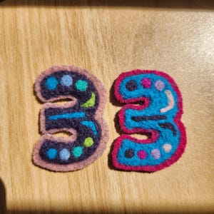May include: Two felt number three patches. The left patch is dark navy blue with a light pink border and features light blue, teal, and lime green polka dots. The right patch is bright blue with a hot pink border and features dark blue, purple, and light blue dots.