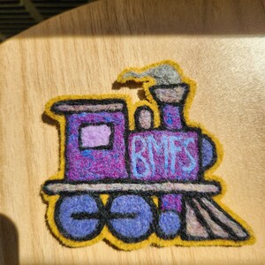 May include: A purple felt train patch with a yellow border, featuring the letters BMFS in light blue on the side. The train has a grey smoke plume, a light purple window, and black outlines defining the wheels, body, and front cowcatcher.