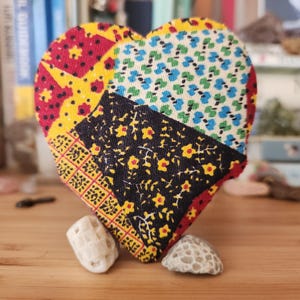May include: A heart-shaped fabric ornament featuring a patchwork design of red, yellow, black, and blue floral and geometric patterns. The heart is propped up by two small, textured, off-white stones that serve as a base for the upright display.