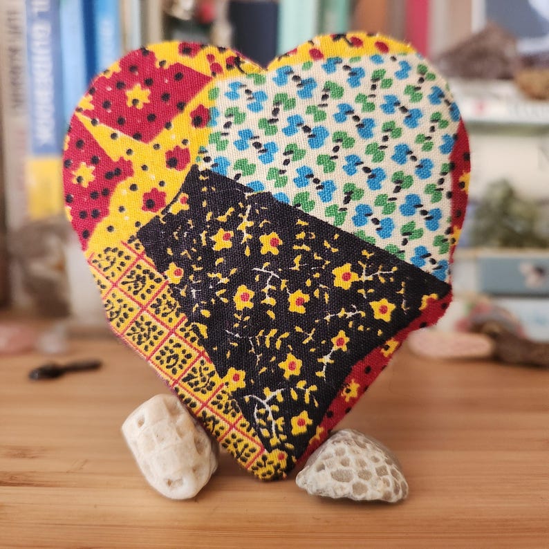 May include: A heart-shaped fabric ornament featuring a patchwork design of red, yellow, black, and blue floral and geometric patterns. The heart is propped up by two small, textured, off-white stones that serve as a base for the upright display.