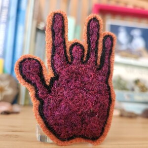 May include: A felted wool patch shaped like a hand making the I love you sign in American Sign Language. The hand is deep magenta with a thick black outline and a bright orange border, featuring a textured, fuzzy surface throughout the design.