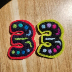 May include: Two felt number three patches. The left patch has a red border and features dark blue felt with green, blue, and light blue circular accents. The right patch has a lime green border and features black felt with blue, purple, and pink accents.