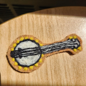 May include: A felt applique shaped like a banjo, featuring a tan outer border with small yellow circular accents, a white circular center, and a black neck with vertical lines. The design is flat and crafted from layered felt material.