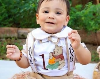 May include: A baby boy wearing a white onesie with a Winnie the Pooh character and the name "ADAR" embroidered on it. He is sitting on a brown and white rug with a red toy helicopter and wooden blocks.