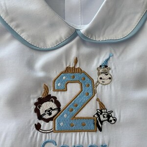 May include: White button-up shirt with a light blue trim on the collar. The shirt features an embroidered number 2 with a blue and gold design, surrounded by animal characters wearing party hats. The name "Conor" is embroidered below.