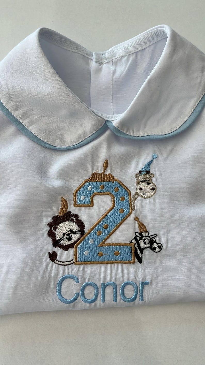 May include: White button-up shirt with a light blue trim on the collar. The shirt features an embroidered number 2 with a blue and gold design, surrounded by animal characters wearing party hats. The name "Conor" is embroidered below.