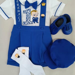 May include: A baby boy's first birthday outfit set. The set includes a white collared shirt with blue trim, blue suspenders, a blue bow tie, and a blue romper. Accessories include blue shoes, a blue cap, and white socks. The outfit features a "Boss Baby" theme.
