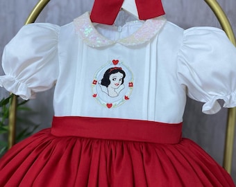 May include: A white and red princess dress with a Snow White embroidered design. The dress features a red bow, a white bodice with puff sleeves, and a red skirt. The collar is embellished with sequins.