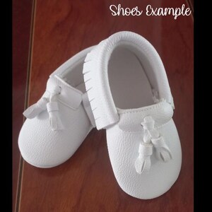 May include: A pair of white baby moccasins with a textured leather appearance. Each shoe features a decorative tassel and fringe detailing. The text "Shoes Example" is written in cursive at the top of the image.