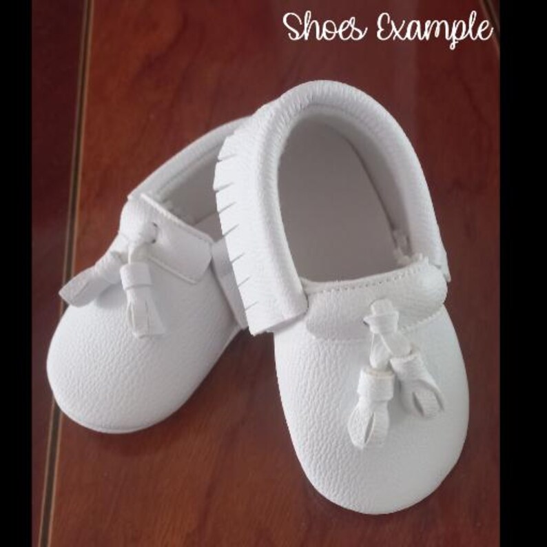 May include: A pair of white baby moccasins with a textured leather appearance. Each shoe features a decorative tassel and fringe detailing. The text "Shoes Example" is written in cursive at the top of the image.