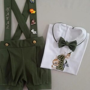 May include: An olive green suspender shorts set with animal-themed embellishments and the name "HAYAZ" embroidered on the suspenders. Includes a white shirt with a green collar, bow tie, and embroidered number "1" with animal designs.