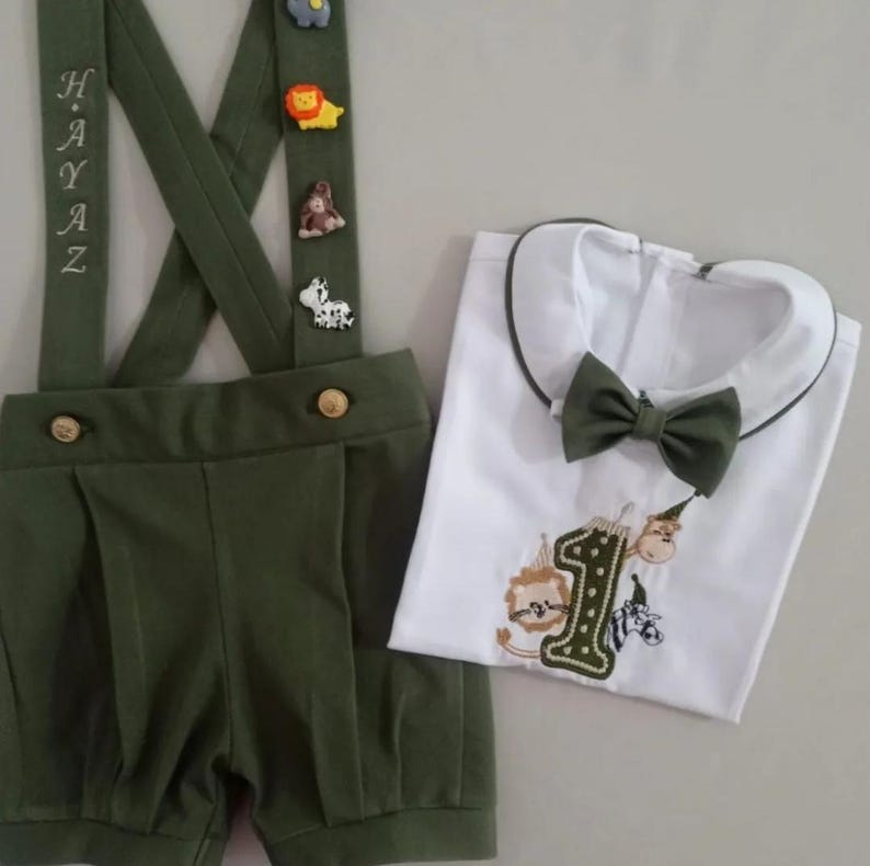 May include: An olive green suspender shorts set with animal-themed embellishments and the name "HAYAZ" embroidered on the suspenders. Includes a white shirt with a green collar, bow tie, and embroidered number "1" with animal designs.