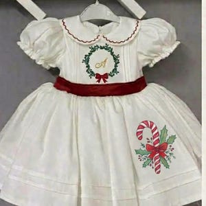 May include: A white dress with puff sleeves, a Peter Pan collar with red trim, and a dark red velvet waistband. The bodice features an embroidered green wreath with a gold letter A, and the skirt displays a candy cane and holly graphic.