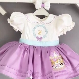 May include: A white dress with a light blue sash and a purple skirt. The dress has a white collar with a scalloped edge and a floral embroidery with the letter "Z" in the center. The skirt has a carousel design with horses and a golden roof.