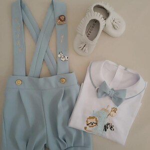 May include: A baby boy's outfit set featuring light blue shorts with suspenders, a white shirt with a bow tie, and white leather shoes. The suspenders have animal-themed embellishments and the shirt has an embroidered design.