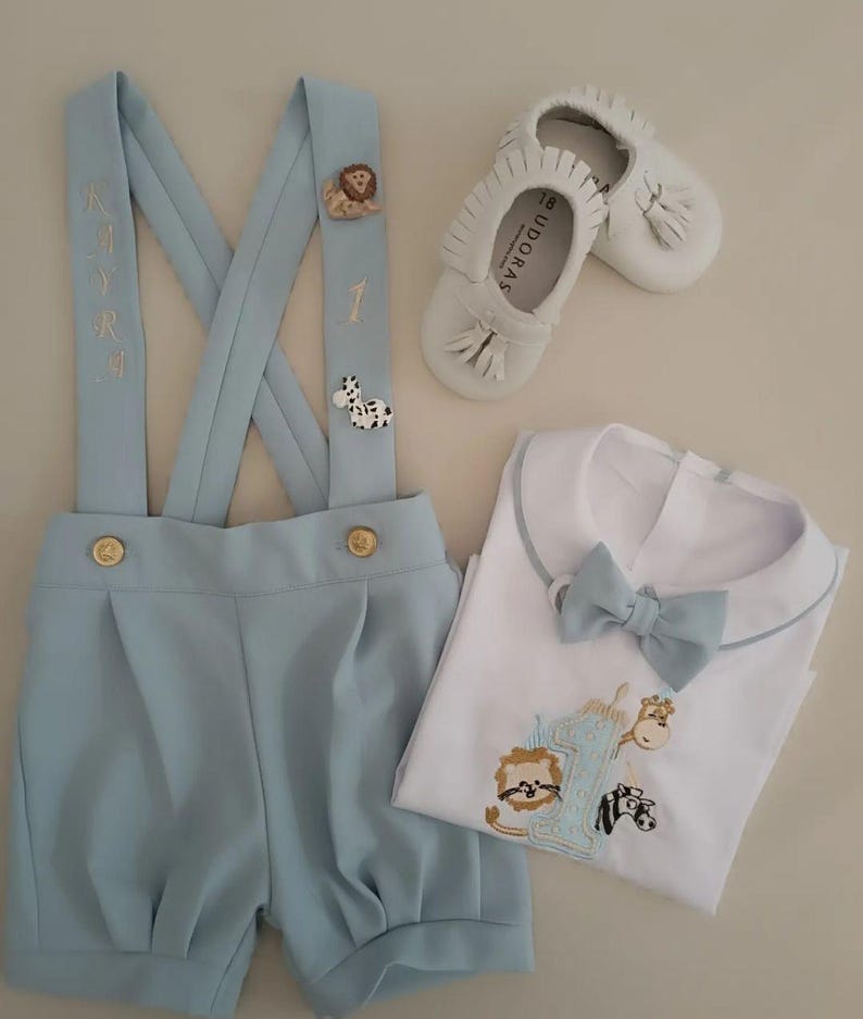 May include: A baby boy's outfit set featuring light blue shorts with suspenders, a white shirt with a bow tie, and white leather shoes. The suspenders have animal-themed embellishments and the shirt has an embroidered design.