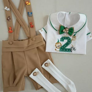 May include: A tan shorts set with suspenders adorned with animal-shaped appliqu&eacute;s. The set includes a white shirt with a green-trimmed collar and a green bow tie. The shirt features an embroidered number 2 and animal designs. White knee-high socks with lion accents complete the outfit.