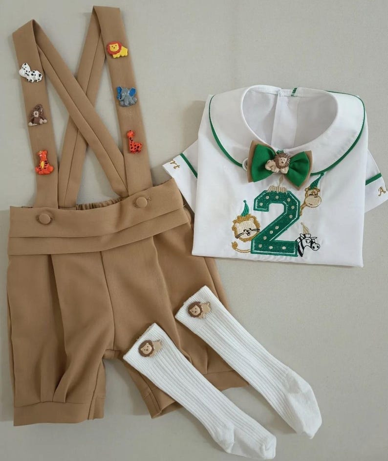 May include: A tan shorts set with suspenders adorned with animal-shaped appliqu&eacute;s. The set includes a white shirt with a green-trimmed collar and a green bow tie. The shirt features an embroidered number 2 and animal designs. White knee-high socks with lion accents complete the outfit.