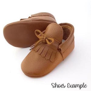 May include: A pair of brown leather baby moccasins with a fringed vamp and a knotted bow detail. The shoes are made of a soft, textured leather and are designed for comfort. The text "Shoes Example" is at the bottom of the image.