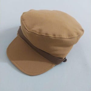 May include: A tan-colored, military-style cap with a short brim. The cap features a brown band that wraps around the crown. The hat is made of a soft fabric and is suitable for various casual outfits.