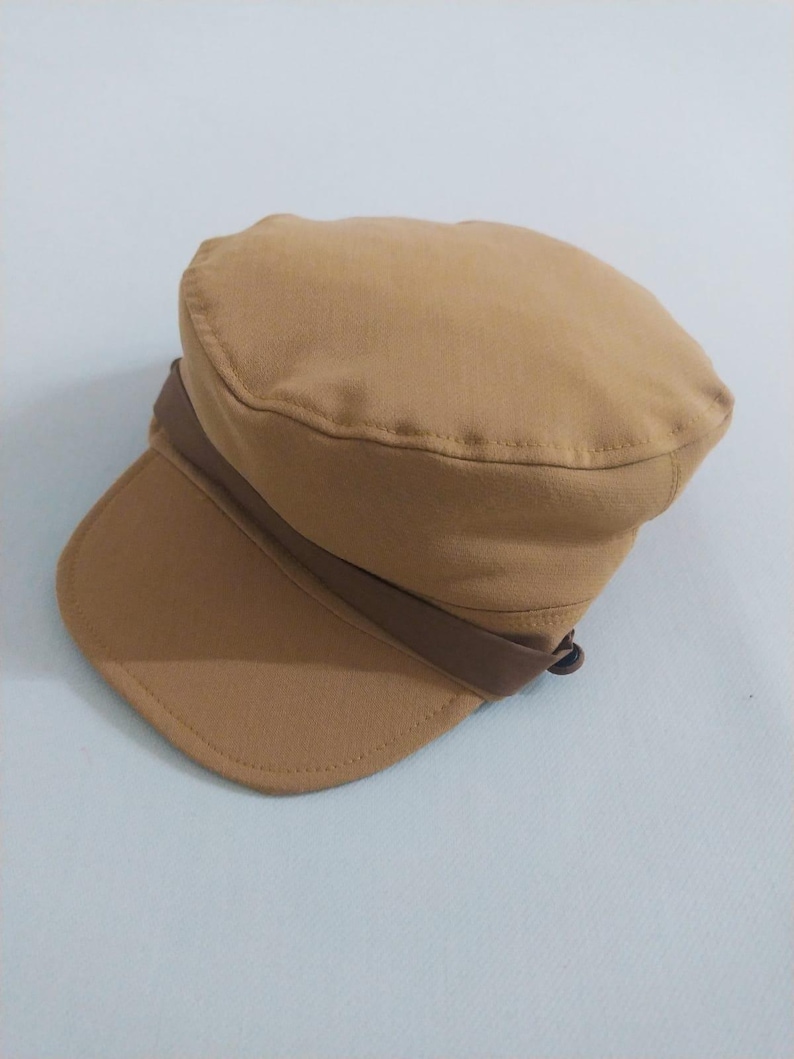May include: A tan-colored, military-style cap with a short brim. The cap features a brown band that wraps around the crown. The hat is made of a soft fabric and is suitable for various casual outfits.
