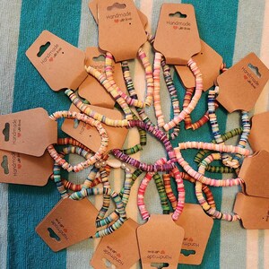 May include: A collection of multi-colored beaded bracelets arranged in a circular pattern. Each bracelet is made of small, flat, circular polymer clay beads in various hues and is attached to a brown cardboard display tag labeled Handmade with love.