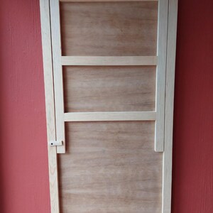 Puede incluir: Una mesa plegable de madera de color claro montada en la pared. La mesa es rectangular con un acabado de veta de madera clara. Cuenta con bisagras de latón y un pestillo para asegurarla en la posición cerrada. La mesa está montada en una pared roja.