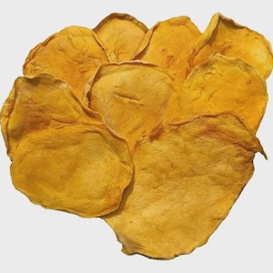 Puede incluir: Un montón de rodajas de mango deshidratado, mostrando un vibrante color amarillo-naranja. Las piezas delgadas, de forma irregular, tienen una textura ligeramente arrugada, lo que sugiere una consistencia masticable. La imagen está sobre un fondo liso y claro, resaltando el color natural de la fruta.