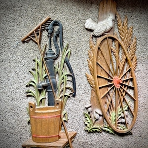 May include: Two rustic wall hangings. One features a water pump, rake, and bucket. The other depicts a wagon wheel, wheat, and a dove. The pieces are made of wood and painted in shades of brown, green, and black.