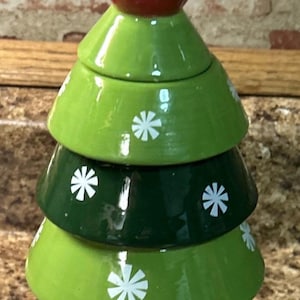 May include: A ceramic Christmas tree-shaped container with a red bird topper wearing a green scarf. The tree is composed of graduated green and dark green sections, each adorned with white snowflake accents.