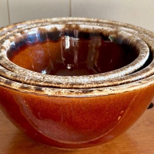 May include: Set of three nested mixing bowls in a warm brown glaze with a lighter rim. The bowls are stacked inside each other, showcasing their graduated sizes. The interior of the bowls is a glossy brown.