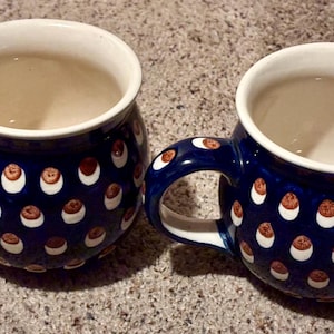 May include: Two ceramic mugs with a dark blue glaze and white interiors. The mugs are decorated with a pattern of white and brown dots. The handles are also blue with the dot pattern. The mugs are sitting on a textured surface.