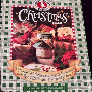 May include: A Gooseberry Patch Christmas Book 3 hardcover book with a green and white checkered border. The cover features a jar of chunky chocolate cookie mix, a red fabric bird, and a banner with the text: Delicious, old-fashioned recipes, handmade holiday gifts & ideas for frosty fun! A Leisure Arts Publication.