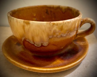 May include: A brown and white ceramic teacup and saucer set with a mottled glaze. The cup has a rounded shape and a handle. The saucer is round and has a slightly raised edge.