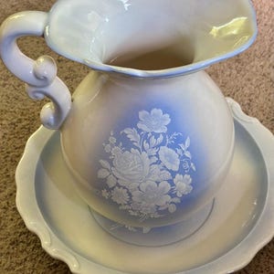 May include: A ceramic pitcher and basin set featuring a white and light blue gradient finish. The pitcher has a curved handle and a scalloped rim, while both pieces are decorated with a white floral pattern of roses and other blossoms.