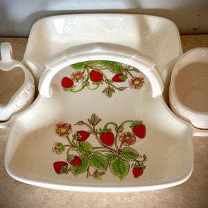 May include: Cream-colored ceramic serving set with a strawberry design. The set includes a basket-shaped tray with a handle, a heart-shaped creamer, and a small bowl. The tray and handle are decorated with red strawberries and green leaves.