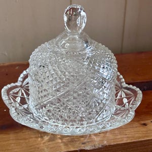 May include: A clear pressed glass butter dish with a dome-shaped lid featuring a diamond point pattern and a faceted knob handle. The base has a scalloped edge with a starburst design, showcasing intricate geometric textures throughout the piece.