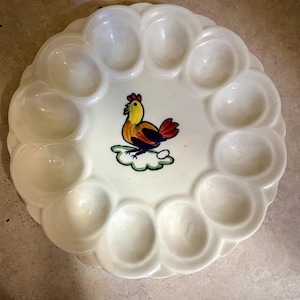 May include: Cream-colored ceramic egg plate with 12 egg indentations. The center features a colorful rooster illustration with yellow, orange, red, and blue hues. The plate has a scalloped edge.
