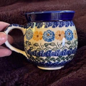 May include: A ceramic mug with a rounded body and a handle. The mug features a blue rim and is decorated with blue, yellow, and green floral patterns. The handle is white. The mug is sitting on a purple surface.
