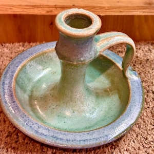 May include: Handmade ceramic candle holder in shades of blue and green. The candle holder has a circular base with a handle and a central cup for the candle. The piece has a rustic, textured appearance, with visible brushstrokes and a glazed finish.