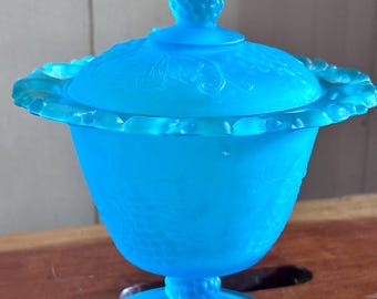 May include: A bright blue satin glass candy dish with a matching lid. The piece features a pedestal base, a scalloped rim, and a textured finial on top. The glass has a frosted finish and is decorated with embossed grape and vine patterns.