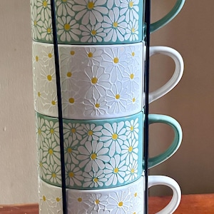May include: A set of four stacked ceramic mugs held in a black metal wire rack. The mugs alternate between a light teal and white background, each featuring a raised, embossed white daisy pattern with yellow centers and matching colored handles.