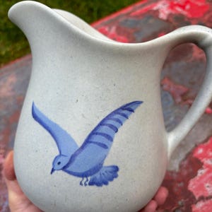 May include: A white ceramic pitcher with a blue bird design. The bird is flying with its wings spread out. The pitcher has a handle and a spout.