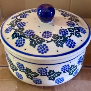 May include: A round ceramic lidded container with a white base and a dark blue knob handle. The exterior features a hand-painted pattern of blue and purple blackberry clusters with dark green leaves, accented by thin blue circular rim lines.