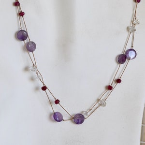 May include: A double-strand necklace with amethyst and ruby beads, and clear crystal accents. The necklace is made of brown cord.