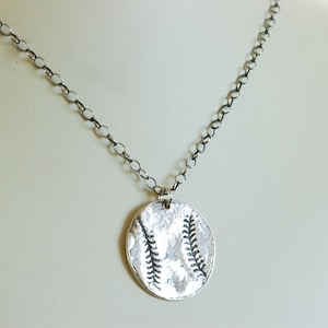Puede incluir: Un collar de cadena de plata con un colgante redondo que presenta un diseño de béisbol. El colgante tiene una superficie texturizada y un diseño de puntadas de béisbol en relieve.