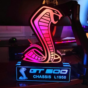 May include: A neon sign featuring a cobra design with pink and white accents. The base of the sign displays the text "GT 500" and "CHASSIS L1958" in blue. The sign is illuminated, creating a vibrant display.