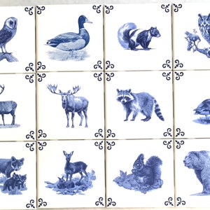 May include: Set of twelve ceramic tiles, each featuring a blue and white illustration of various animals. The collection includes an owl, duck, skunk, deer, moose, raccoon, goose, fox, deer, squirrel, and bear. Each tile has a decorative corner design.