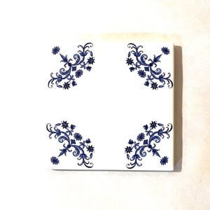 May include: Square white tile with a blue floral design in each corner. The design features stylized flowers and leaves. The tile is likely ceramic and suitable for home decor projects.